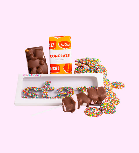A selection of chocolate candies from a gift box, including shapes with messages, milk chocolate pieces, and freckle grab bag, arranged on a white surface with sprinkles and decorative packaging.