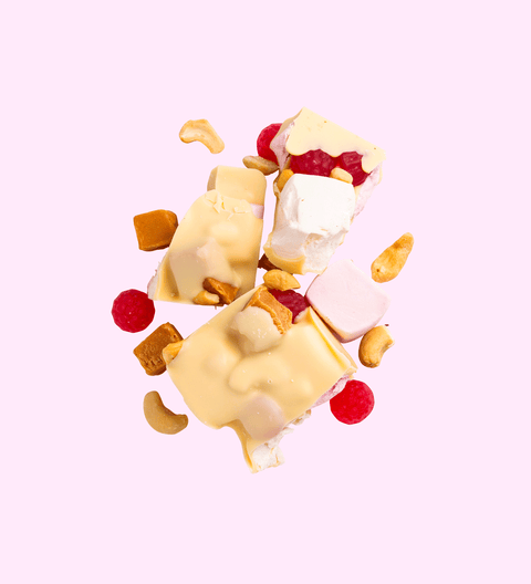 Chunks of white chocolate with visible pieces of marshmallows, cashews, and raspberry lollies scattered against a white background.