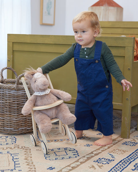 Child in blue overalls pushing a teddy bear in a wooden chair on a patterned rug.