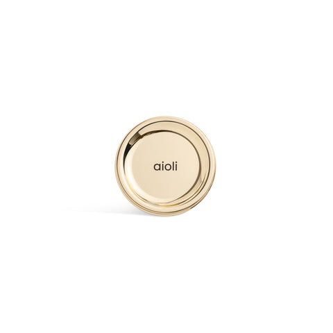A tall, cylindrical white bottle with a brushed gold label disc at the bottom and a precision squeeze nozzle on the side. Aioli sauce