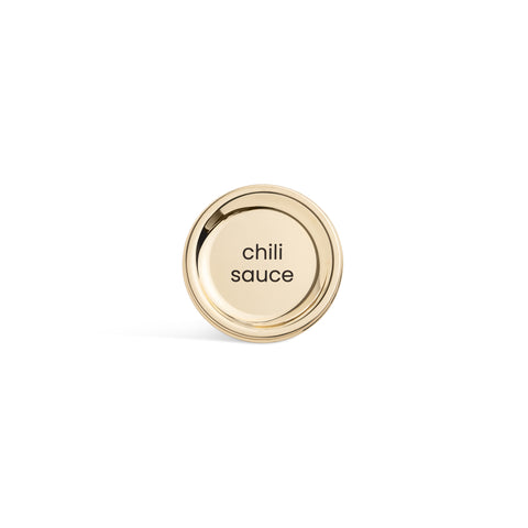 A tall, cylindrical white bottle with a brushed gold label disc at the bottom and a precision squeeze nozzle on the side. Chili sauce