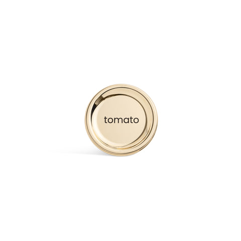 A tall, cylindrical white bottle with a brushed gold label disc at the bottom and a precision squeeze nozzle on the side. Tomato