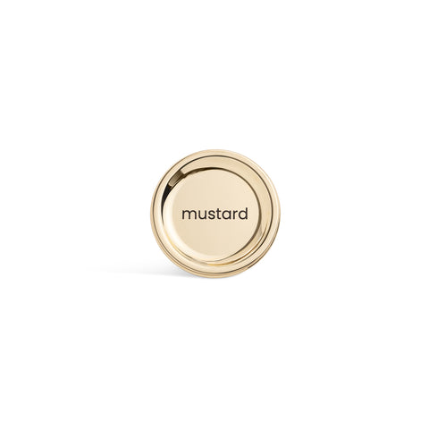 A tall, cylindrical white bottle with a brushed gold label disc at the bottom and a precision squeeze nozzle on the side. Mustard
