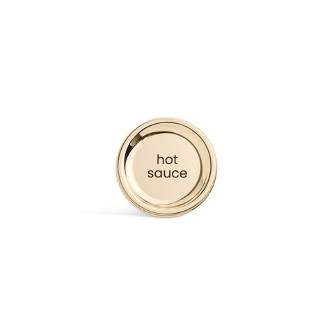 A tall, cylindrical white bottle with a brushed gold label disc at the bottom and a precision squeeze nozzle on the side. Hot Sauce