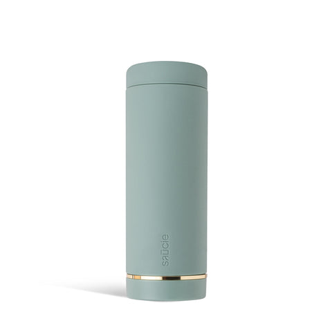 A tall, cylindrical sage green Saucie bottle with a brushed gold label disc at the bottom.