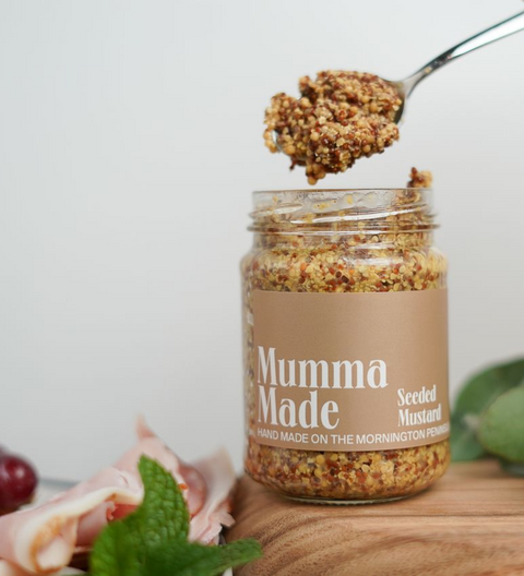 Jar of 'Mumma Made' seeded mustard with a spoonful above, on a wooden surface.