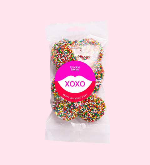 A clear plastic bag filled with multicolored chocolate freckles, sealed with a pink label that reads 'Happy Valentine's Day'.