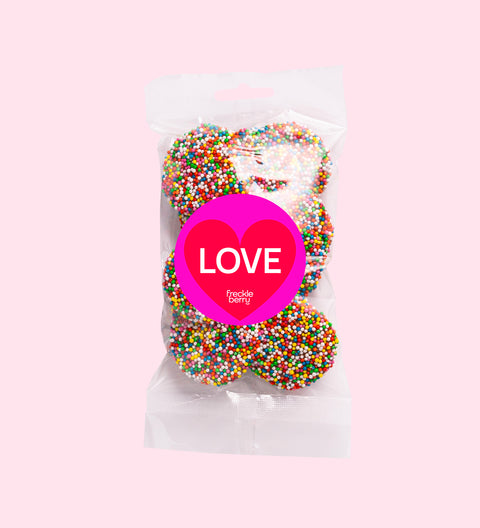 A clear plastic bag filled with multicolored chocolate freckles, sealed with a pink label that reads 'Happy Valentine's Day'.