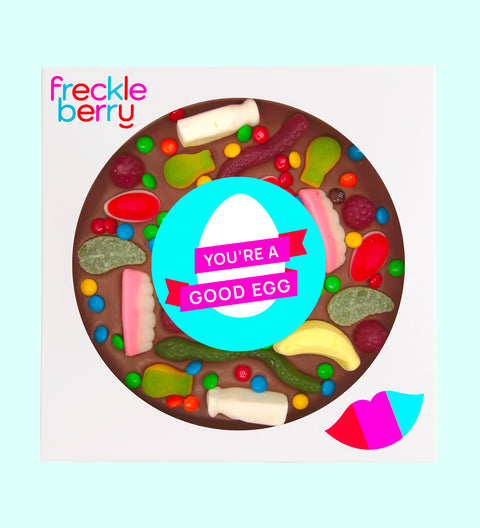 A giant chocolate pizza with various lolly toppings, including chocolate buttons and candies, with a text message in the center that reads 'YOU'RE A GOOD EGG'. The pizza is circular and has a 18cm diameter, with the product branding 'freckleberry' at the top.