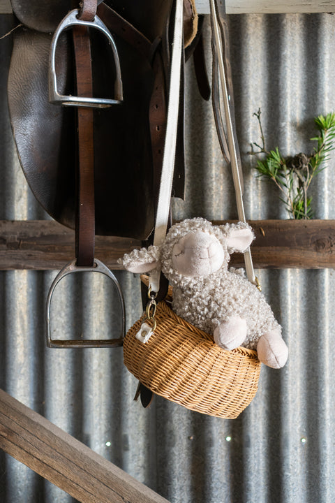 Sheep plush toy in a woven basket hanging on a metal wall with a saddle and stirrup.