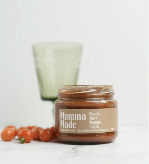 Jar of 'Mumma Made' relish with tomatoes and a glass in the background on a white surface.