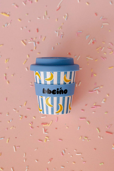 A reusable coffee cup with a blue lid and blue sleeve, decorated with blue and white vertical stripes and yellow banana illustrations, set against a pink background with scattered colorful sprinkles. The brand name 'bbció' is printed on the sleeve.