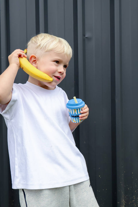 Child holding a banana and a blue cup with a straw against a dark background