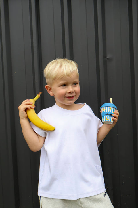 A young blonde boy wearing a white t-shirt and light gray sweatpants stands in front of a dark gray corrugated metal wall, holding a yellow banana in one hand and a blue striped cup with a straw in the other.