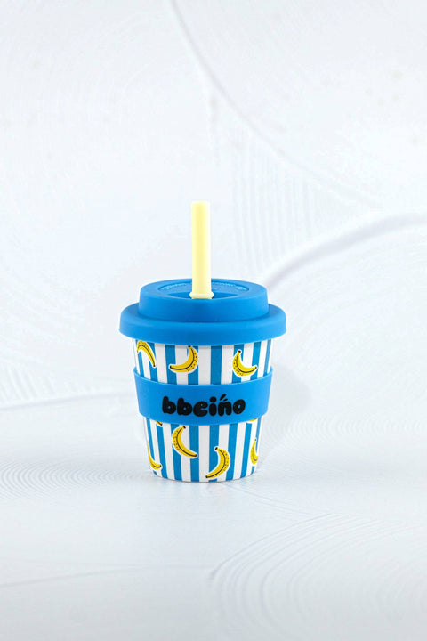 A small novelty cup-shaped accessory with a blue lid and straw, blue-and-white vertical stripes, yellow banana illustrations, and a blue band reading 'bbciño' in black text, shown on a light gray background.