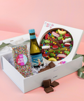 Gift box with a bottle of Prosecco, a colorful chocolate pizza, and a packet of sprinkles on a pink background.