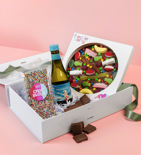 Gift box with a bottle of Prosecco, a colorful chocolate pizza, and a packet of sprinkles on a pink background.