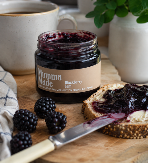 An open jar of blackberry jam labeled 'Mumma Made' sits on a wooden surface beside a slice of bread spread with jam, blackberries, a knife, a mug, and a potted plant in a kitchen setting.