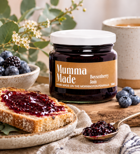 A jar of Mumma Made Boysenberry Jam is shown on a wooden table with a slice of toast spread with jam, blueberries, a spoonful of jam, and a mug in the background, creating a cozy breakfast scene.