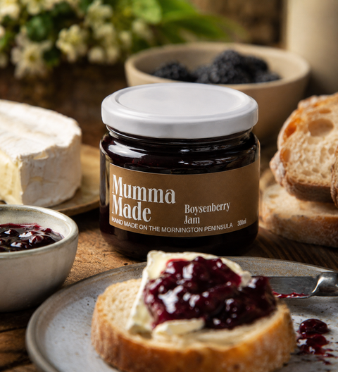 A jar of Mumma Made Boysenberry Jam is shown on a rustic table with bread, cheese, and a small bowl of jam. The jar has a white lid and a brown label with white text.