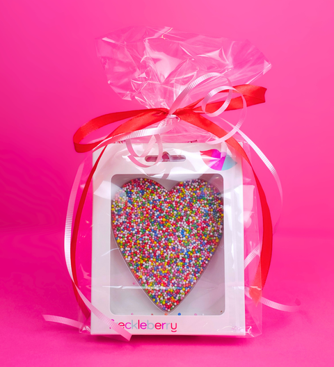 Heart-shaped decorative item with colorful sprinkles in a clear gift box with a red ribbon on a pink background.