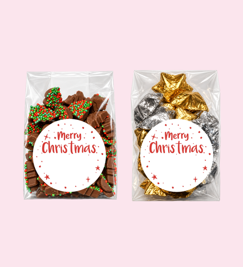 Two clear gift bags filled with holiday treats on a pale pink background. The left bag contains assorted chocolate pieces with red and green sprinkles, and the right bag contains foil-wrapped chocolates in gold and silver.