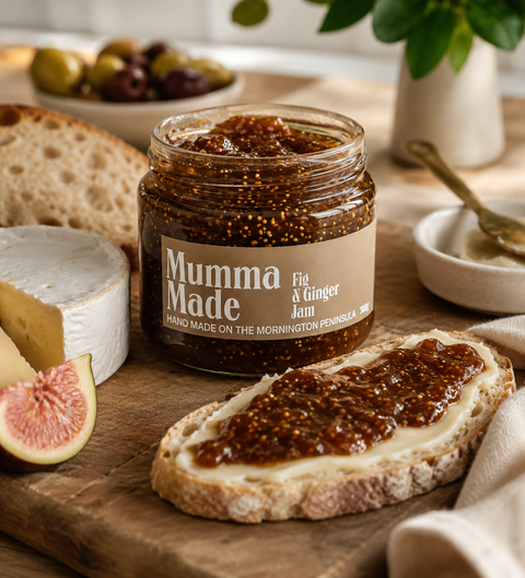 A jar of Mumma Made Fig & Ginger jam sits on a wooden board with bread, cheese, figs, and a spoon in a softly lit kitchen setting.