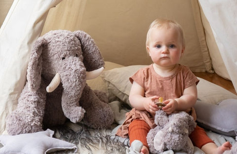 Baby sitting on a bed with a large plush elephant toy next to them