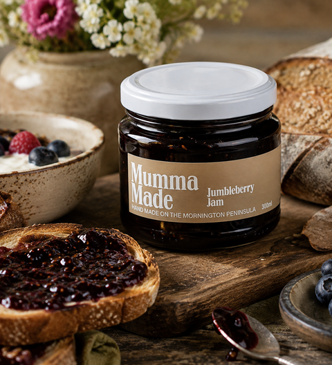 A jar of Mumma Made Jumbleberry Jam is displayed on a rustic wooden table surrounded by food props, including a slice of toast with jam, blueberries, bread, and a bowl of yogurt with berries.
