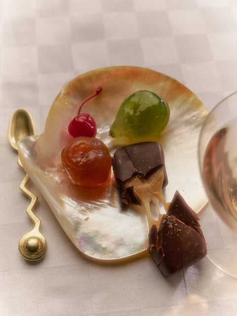 A small shell-shaped dish holding assorted sweets and chocolates on a checkered tablecloth, with a gold spoon and part of a glass visible.