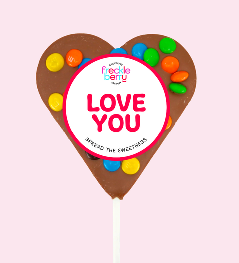 Heart-shaped chocolate lollipop with colorful candies and 'Love You' branding on a pink background.