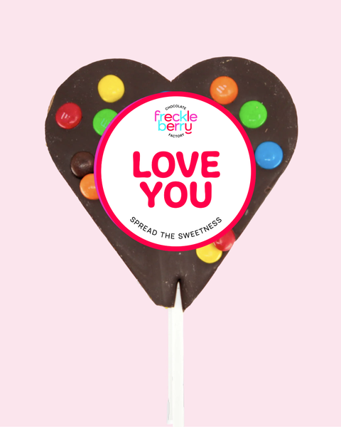 Heart-shaped chocolate lollipop with colorful candies and 'Freckle Berry' branding on a pink background.