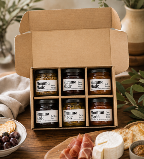 An open cardboard gift box containing six jars of Mumma Made spreads, styled on a wooden table with cheese, crackers, olives, and prosciutto.