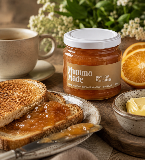 A jar of Mumma Made Breakfast Marmalade stands on a wooden surface beside toast spread with jam, butter, sliced orange, and a cup, with flowers blurred in the background.