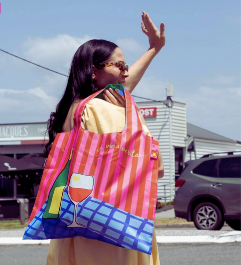 Woman holding a colorful striped bag with a store and car in the background. My Shout Reusable Shopping Bag