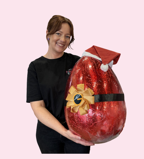 Person holding a large 6kg red chocolate Easter egg with a Santa hat and bow on a pink background