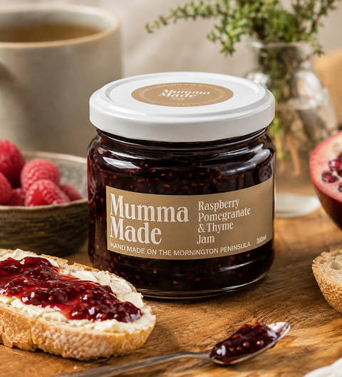 A jar of Mumma Made Raspberry Pomegranate & Thyme Jam sits on a wooden surface with a slice of toast spread with jam in the foreground and fresh raspberries nearby, creating a cozy, rustic lifestyle scene.