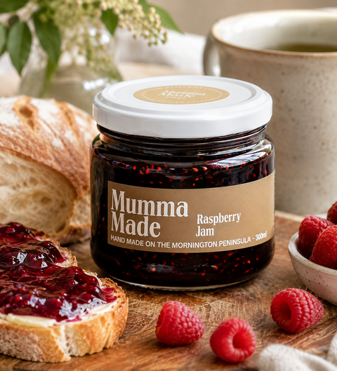 A jar of Mumma Made Raspberry Jam sits on a wooden surface surrounded by food props, including a slice of bread with jam spread, fresh raspberries, and a ceramic mug in the background.