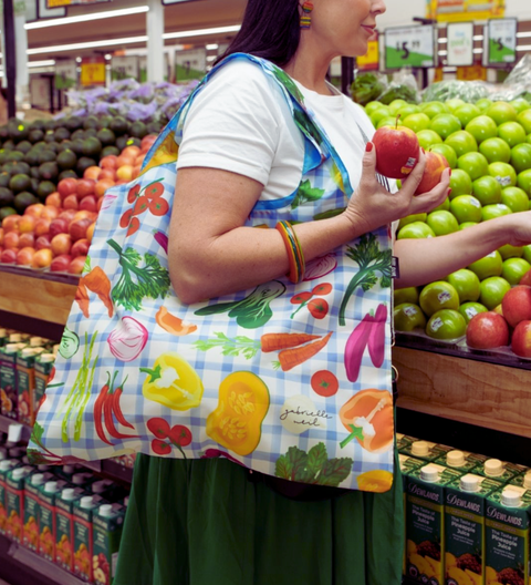 Woman in a grocery store holding apples, wearing a colorful apron with fruit and vegetable patterns. Harvest Party Reusable Shopping Bag Ruby Olive - Reusable shopping bag with vegetable pattern on a blue checkered background