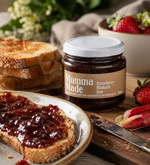 A jar of Mumma Made Strawberry Rhubarb Jam is shown on a rustic wooden table with toast, strawberries, and rhubarb, creating a cozy breakfast scene.