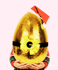 Person holding a large gold Easter egg with a red bow and Santa hat on a white background