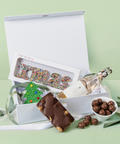 Christmas Gift box with assorted treats including a bottle, candy, and chocolate bar on a light green background.