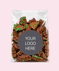 A clear plastic gift bag filled with assorted chocolate pieces topped with red and green holiday sprinkles, centered on a light pink background. A dark gray oval label in the middle reads 'YOUR LOGO HERE'.
