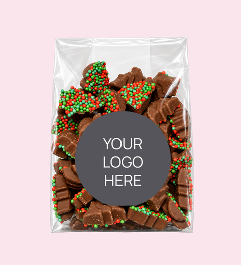 A clear plastic gift bag filled with assorted chocolate pieces topped with red and green holiday sprinkles, centered on a light pink background. A dark gray oval label in the middle reads 'YOUR LOGO HERE'.
