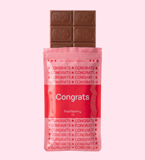 A chocolate bar in a bright pink wrapper labeled 'Congrats' and 'freckleberry', with a dark brown chocolate bar partially pulled from the top, on a solid pale pink background.