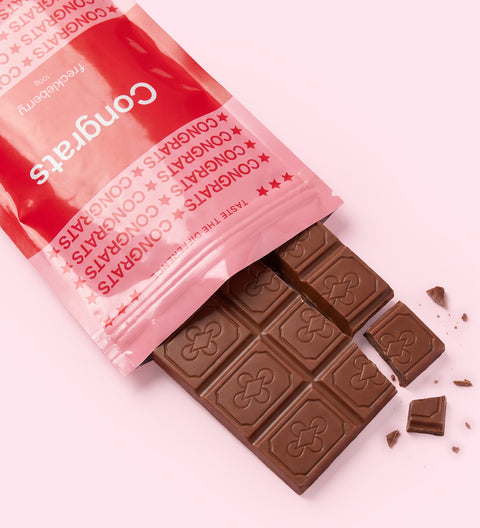A pink and red 'Congrats' chocolate bar wrapper with a broken chocolate bar and crumbs on a light pink background.