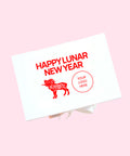 A white rectangular gift box or card package shown at an angle on a light pink background. The front has red text reading 'HAPPY LUNAR NEW YEAR,' a red ram illustration with floral patterning, and a red circular placeholder that says 'YOUR LOGO HERE.