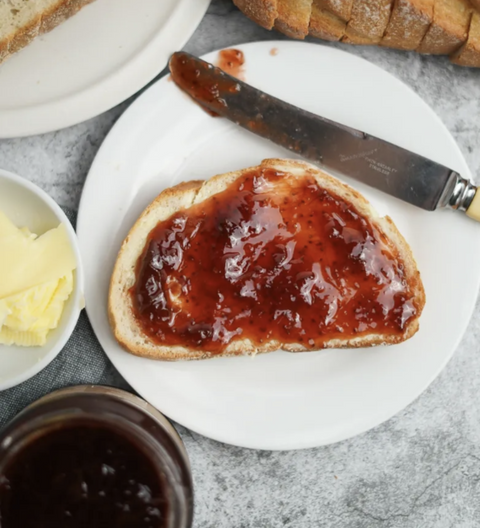 Toast with strawberry jam on a white plate, surrounded by a jar of jam, a knife, and a small bowl of butter.