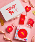 A styled flat-lay of Lunar New Year themed gift items on a pink background, including a white gift box with red text and a red animal graphic, a red heart-shaped lollipop, a red rectangular item, a small bag of red candies, and a round red item on a white tin labeled 'frecklberry'.
