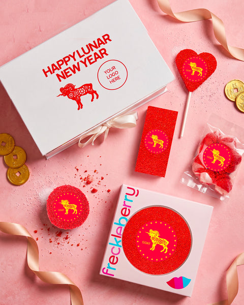 A styled flat-lay of Lunar New Year themed gift items on a pink background, including a white gift box with red text and a red animal graphic, a red heart-shaped lollipop, a red rectangular item, a small bag of red candies, and a round red item on a white tin labeled 'frecklberry'.